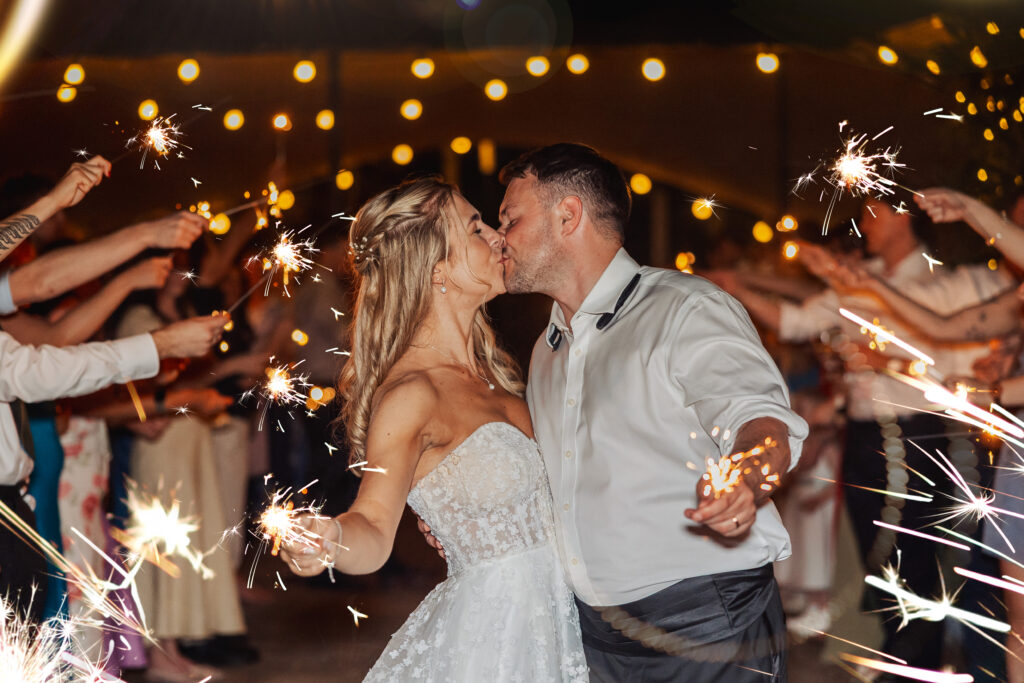 A bride and groom kiss while holding sparklers, surrounded by guests also holding sparklers, under string lights at a nighttime wedding celebration.