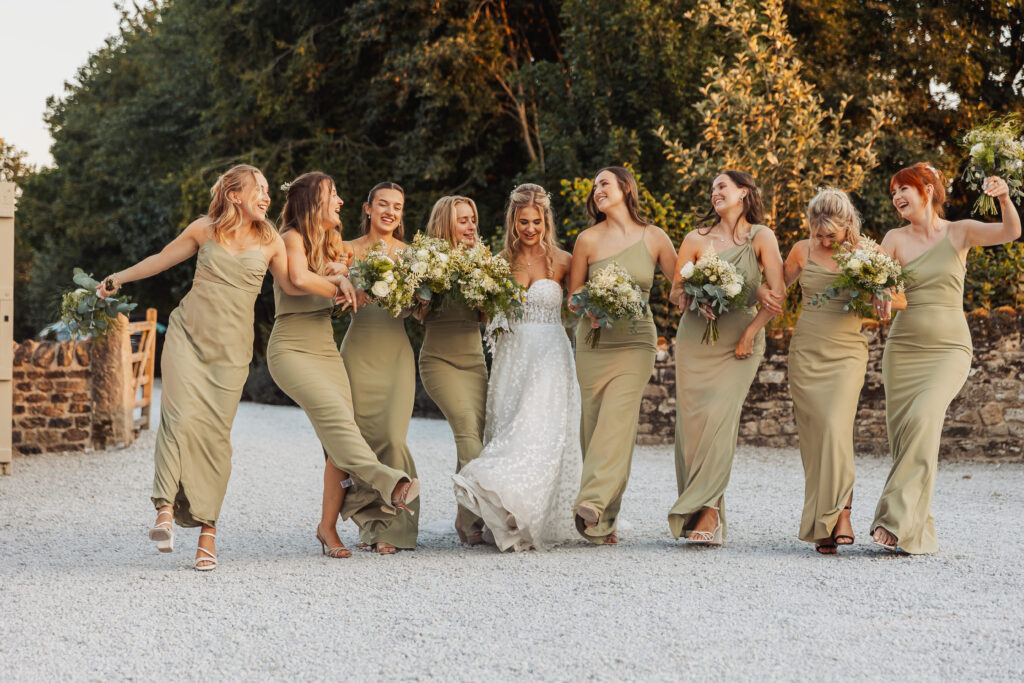 A bride in a white gown walks outdoors with seven bridesmaids in matching sage green dresses, all smiling and carrying bouquets, surrounded by trees and stone walls at a picturesque North Yorkshire wedding.
