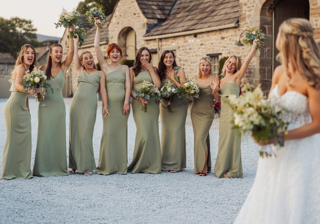 A group of bridesmaids in sage green dresses stand outdoors at Bolton Abbey, smiling and cheering as they see the bride, who is in the foreground with her back to the camera, bouquet in hand. Stone buildings complete this North Yorkshire wedding scene.