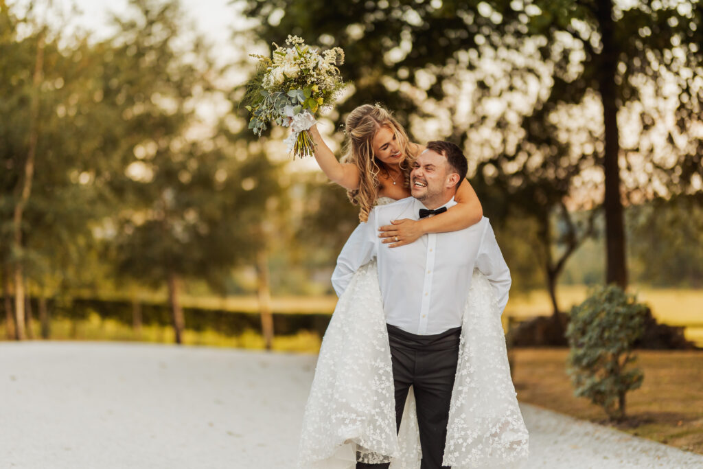 A bride in a white dress holds a bouquet and rides piggyback on the groom, both smiling and joyful, along a sunlit path at their Bolton Abbey wedding, surrounded by trees.
