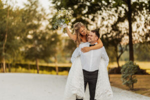 A bride in a white dress holds a bouquet and rides piggyback on the groom, both smiling and joyful, along a sunlit path at their Bolton Abbey wedding, surrounded by trees.