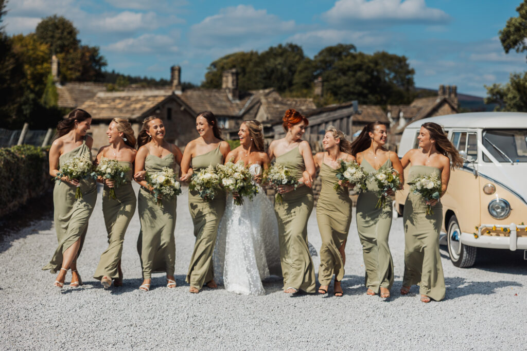A bride in a white dress walks outdoors with eight bridesmaids in sage green, all smiling and holding bouquets, with a vintage cream van and stone buildings in the background at a beautiful North Yorkshire wedding near Bolton Abbey.