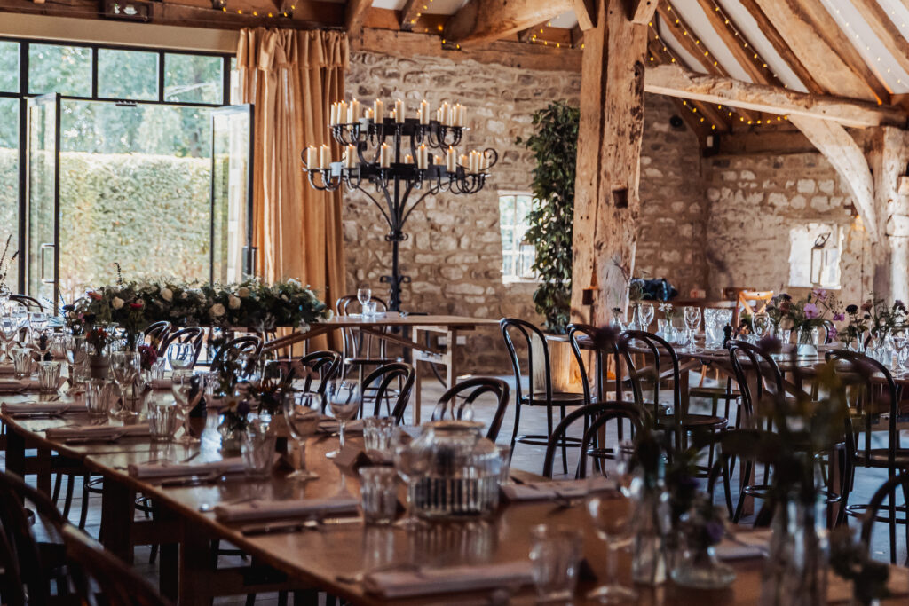 A rustic, elegant dining room in a tithe barn with stone walls and wooden beams, set for a North Yorkshire wedding. Long tables are adorned with glassware, flowers, and candles; sunlight pours through large windows beside a candelabra.