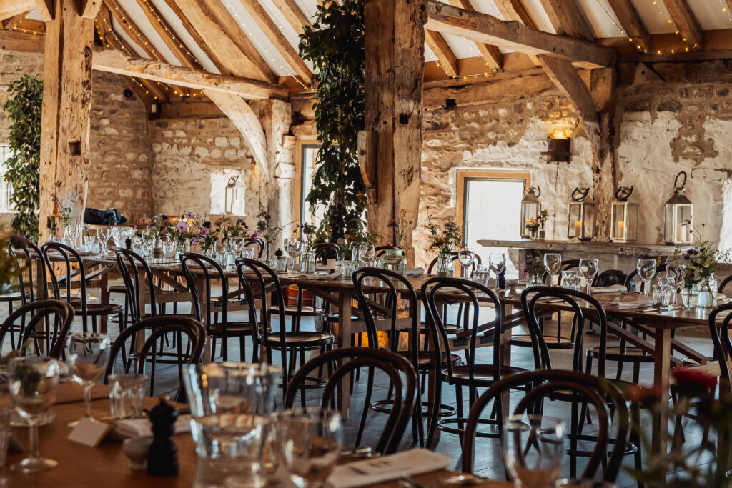A rustic banquet hall at Tithe Barn near Bolton Abbey, with wooden beams, stone walls, and string lights, set for a North Yorkshire wedding—rows of tables and chairs adorned with flowers and glassware, ready for celebration.