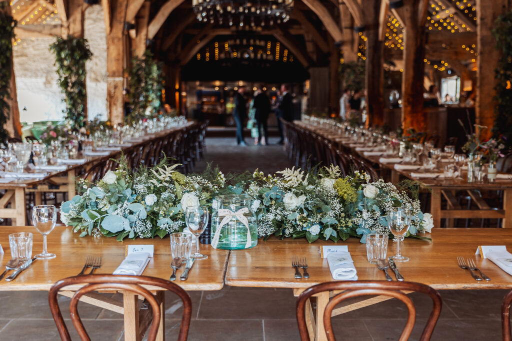A rustic banquet hall at Bolton Abbey with long wooden tables set for a formal wedding, adorned with lush green and white floral centerpieces, glassware, and place settings, under warm string lights and exposed wooden beams.