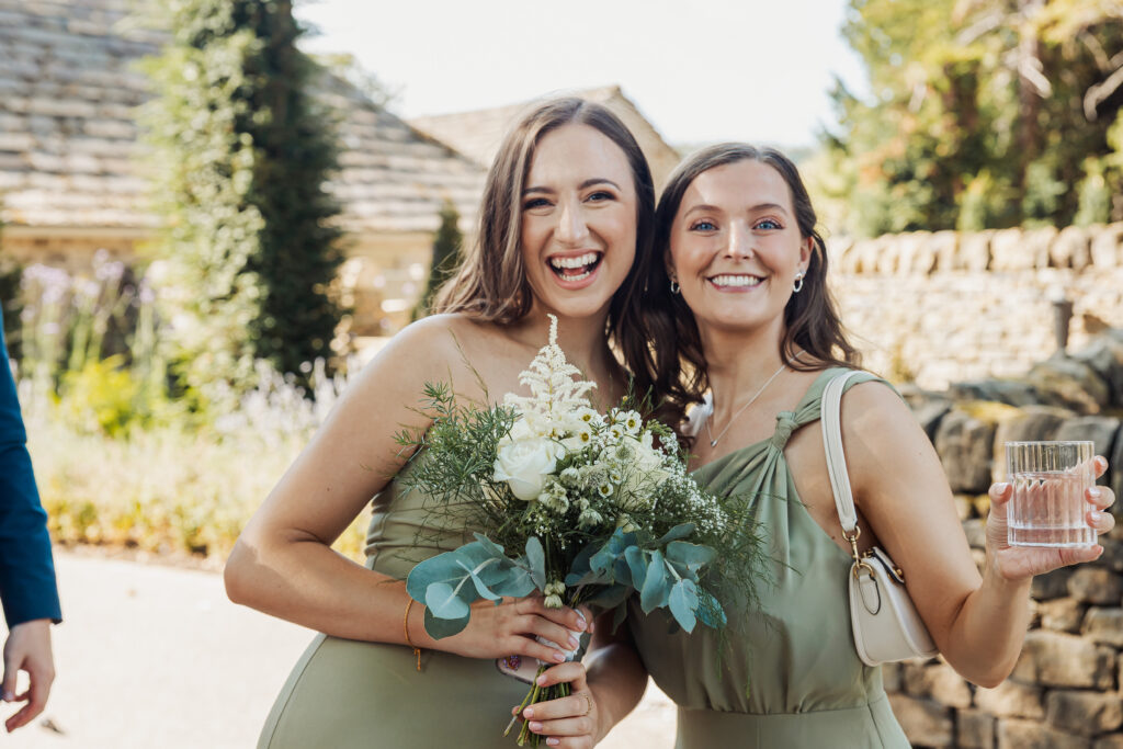Two women in light green dresses smile at the camera outdoors at Tithe Barn, Bolton Abbey. One holds a bouquet of white flowers and greenery, the other a glass of water. A stone wall and lush greenery set a picturesque wedding backdrop.