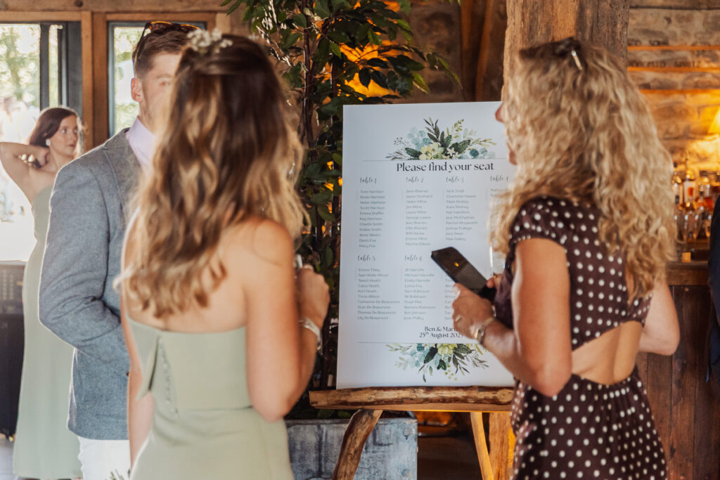 Two women and a man stand indoors at a North Yorkshire wedding, looking at a large seating chart decorated with greenery on an easel as sunlight streams through the window—capturing a charming moment at the Tithe Barn near Bolton Abbey.