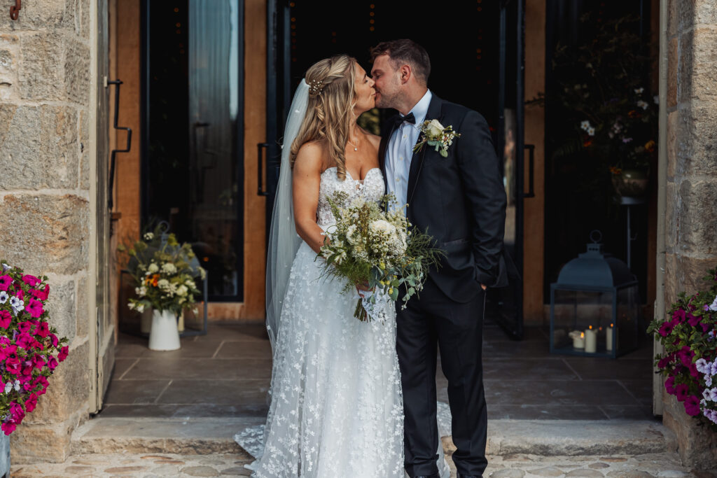 A bride and groom stand close together, smiling and touching noses outside a building at Bolton Abbey. The bride holds a bouquet of white flowers, and both wear formal wedding attire with floral decorations for their North Yorkshire wedding.