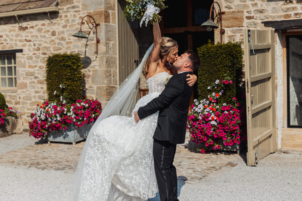 A groom in a black suit lifts the bride, who wears a white dress and holds a bouquet overhead, as they smile and kiss outside Tithe Barn at their North Yorkshire wedding, framed by a rustic stone building adorned with bright flowers.