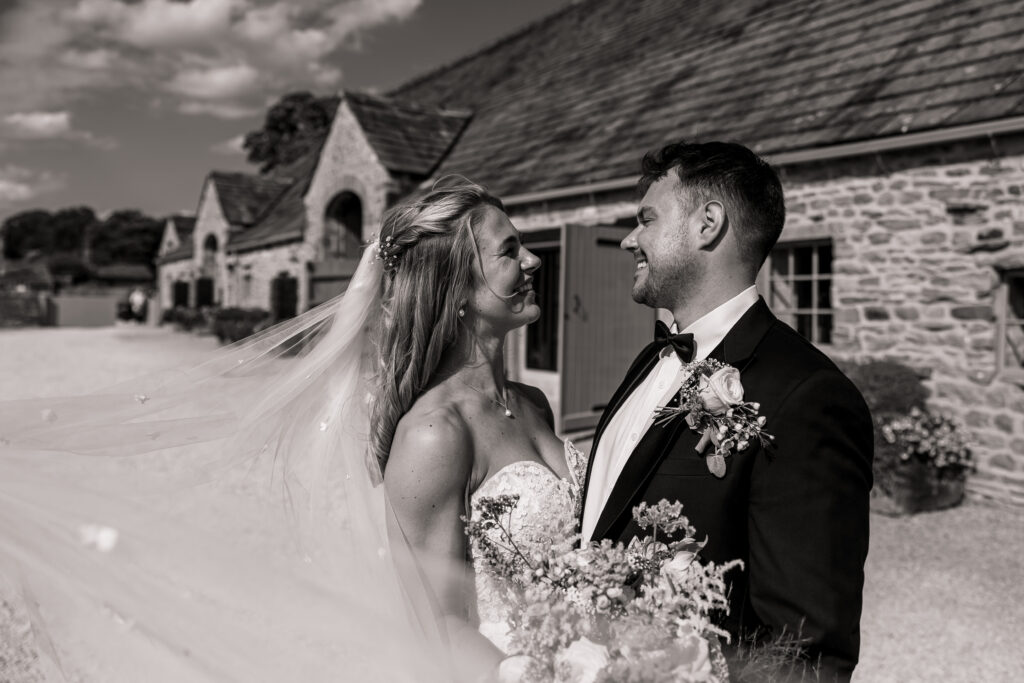 A bride and groom stand outside the historic Tithe Barn in North Yorkshire, smiling at each other happily. The bride wears a strapless gown with a veil, and the groom sports a suit with boutonniere on this sunny wedding day.