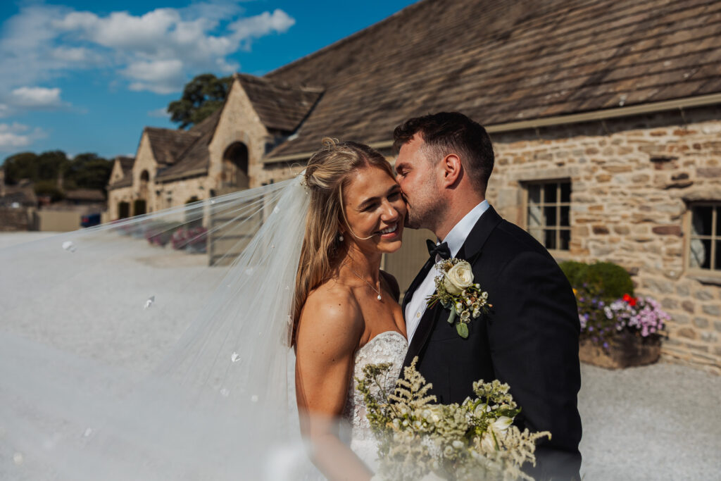 A groom in a tuxedo kisses his smiling bride on the cheek outside a rustic stone building at Bolton Abbey, her veil flowing and bouquet in hand, capturing the joy of their North Yorkshire wedding on a sunny day.