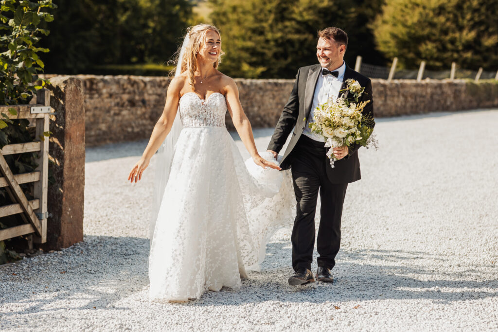 A bride in a white gown and a groom in a tuxedo walk hand in hand outdoors, smiling joyfully. At their North Yorkshire wedding, she holds her dress while he carries a bouquet as they stroll on a sunlit gravel path.