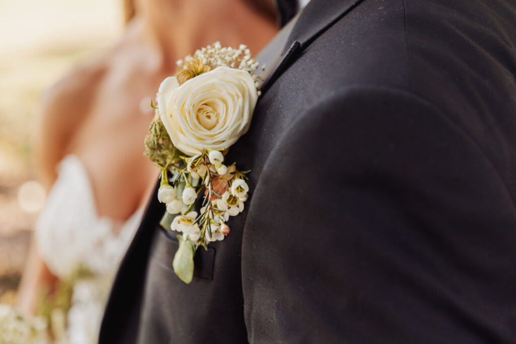 Close-up of a person in a black suit wearing a boutonniere with a white rose and small flowers at a north Yorkshire wedding. Another person in a white, lace wedding dress stands beside them, slightly out of focus.