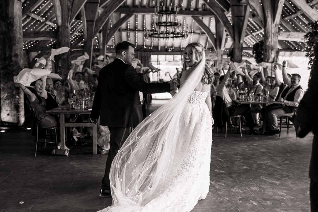 A bride and groom, holding hands and smiling, walk through a rustic North Yorkshire wedding banquet hall near Bolton Abbey as guests cheer and wave napkins. The bride’s long veil trails behind her in this joyful, celebratory scene.