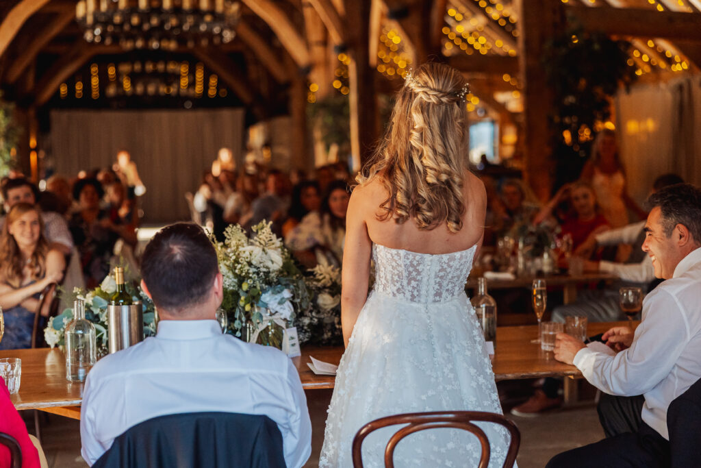 A bride in a white wedding dress stands facing seated guests in a warmly lit tithe barn, decorated with fairy lights, delivering a speech at a long table adorned with flowers, bottles, and glasses at her North Yorkshire wedding.