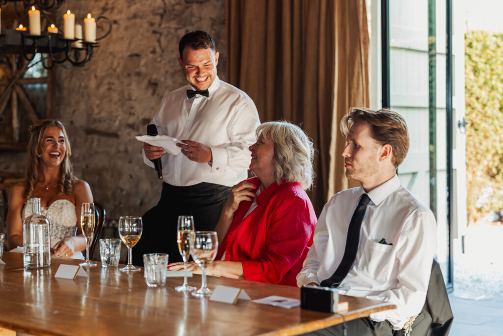 A man in a tuxedo stands and smiles, holding a microphone and speech paper, as three guests laugh at a wooden table set with drinks in the warmly lit Tithe Barn at Bolton Abbey during a joyful wedding celebration.