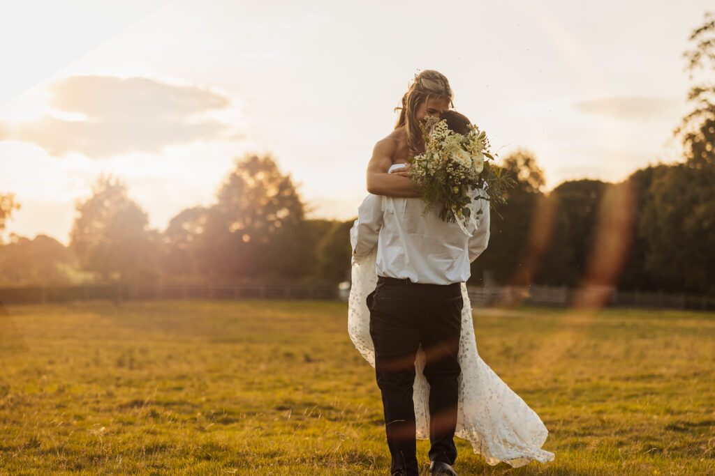 A person in a white dress is lifted by another in a white shirt and dark pants in a sunlit field at a North Yorkshire wedding, holding a bouquet of flowers, with trees and a golden sky in the background.
