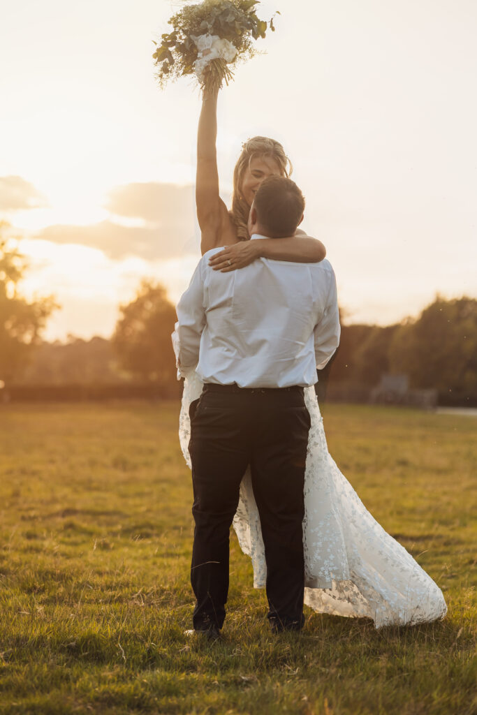 A bride in a white dress is lifted by her groom in a grassy field at sunset near Bolton Abbey, holding her bouquet triumphantly above her head—capturing pure wedding joy.