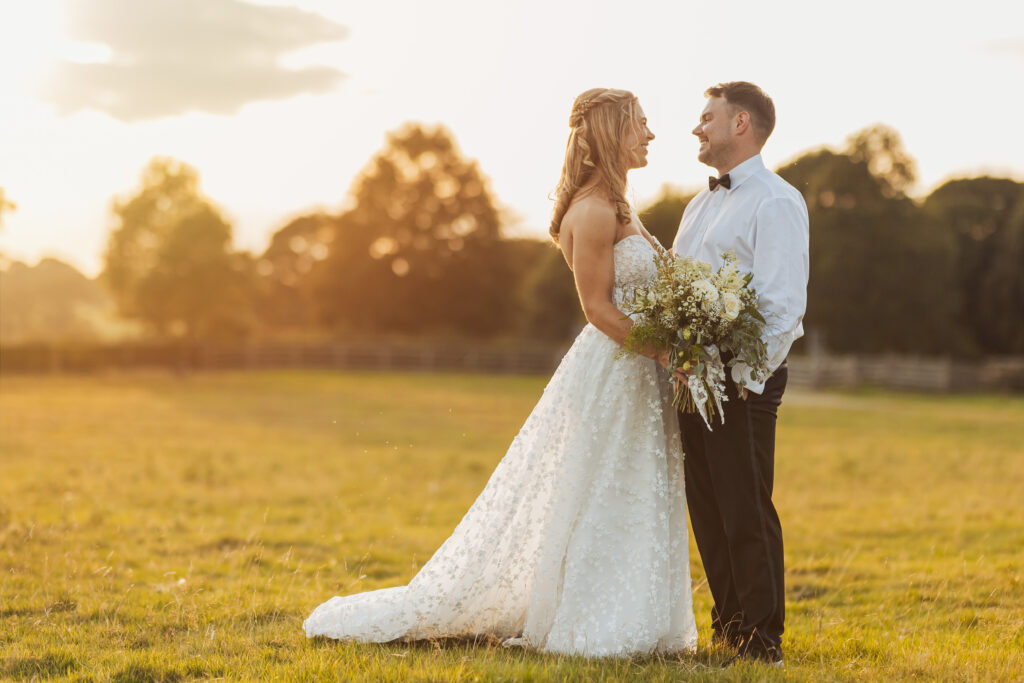 A bride in a white dress and a groom in a white shirt and black pants stand facing each other in a sunlit field, holding a bouquet and smiling, at their North Yorkshire wedding with trees and a sunset in the background.