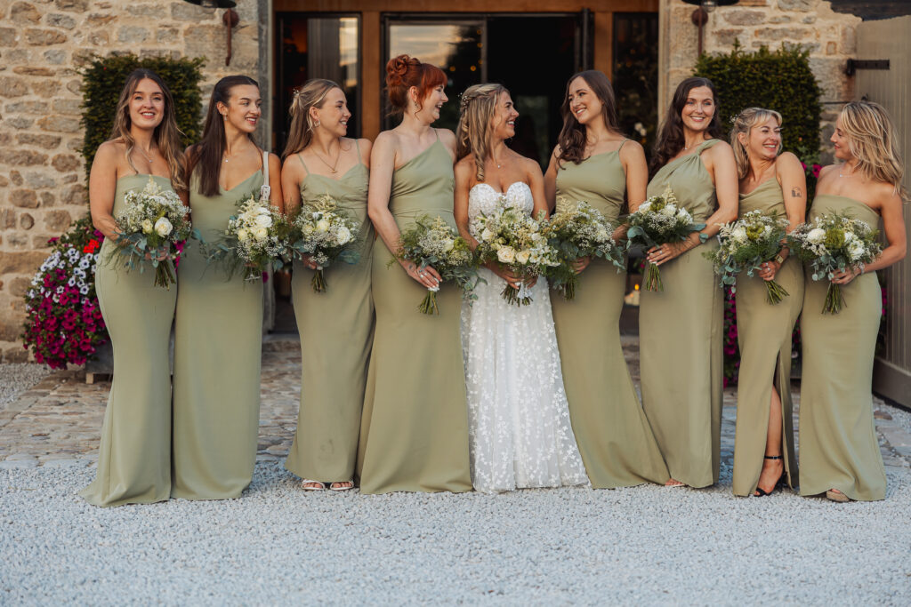 A bride in a white dress stands with seven bridesmaids in matching sage green dresses, all holding bouquets and smiling together outside the Tithe Barn at a beautiful North Yorkshire wedding.