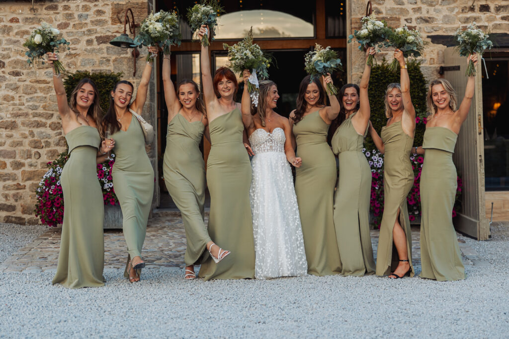 A bride in a strapless white gown stands with eight bridesmaids in matching sage green dresses, all smiling and holding bouquets, celebrating outdoors at a North Yorkshire wedding near the historic Tithe Barn at Bolton Abbey.