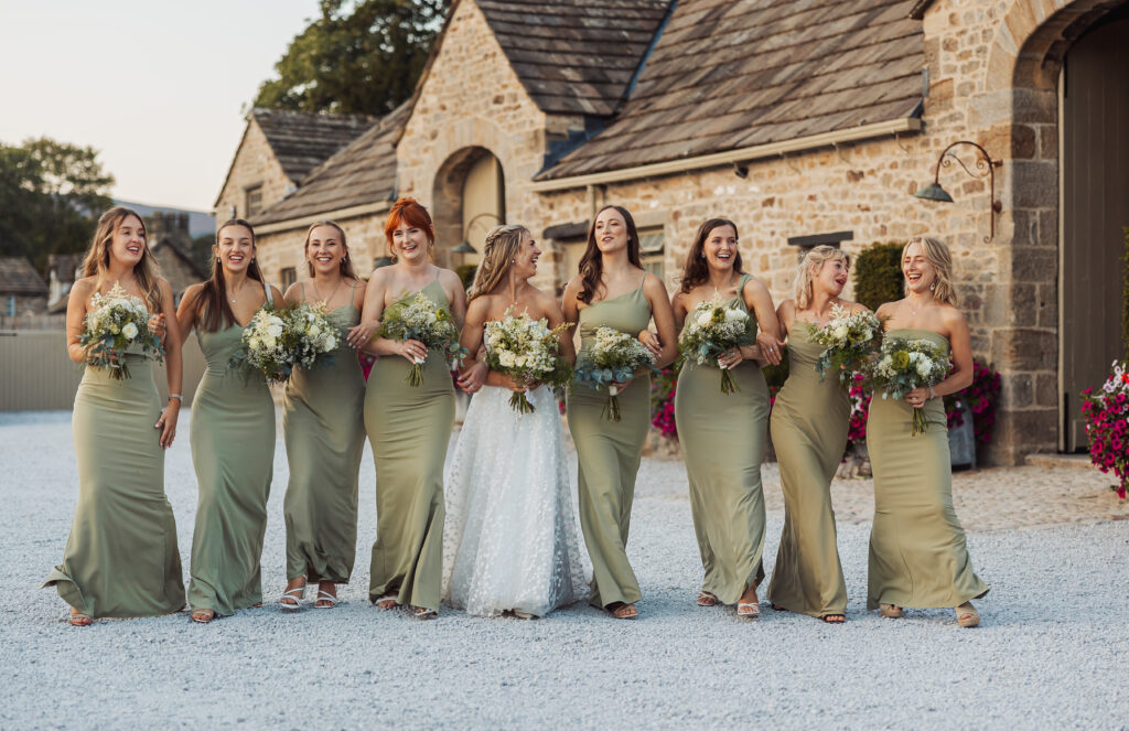 A bride in a white gown walks outside with eight bridesmaids in matching sage green dresses, all holding bouquets and smiling in front of rustic stone buildings at a beautiful Bolton Abbey wedding.