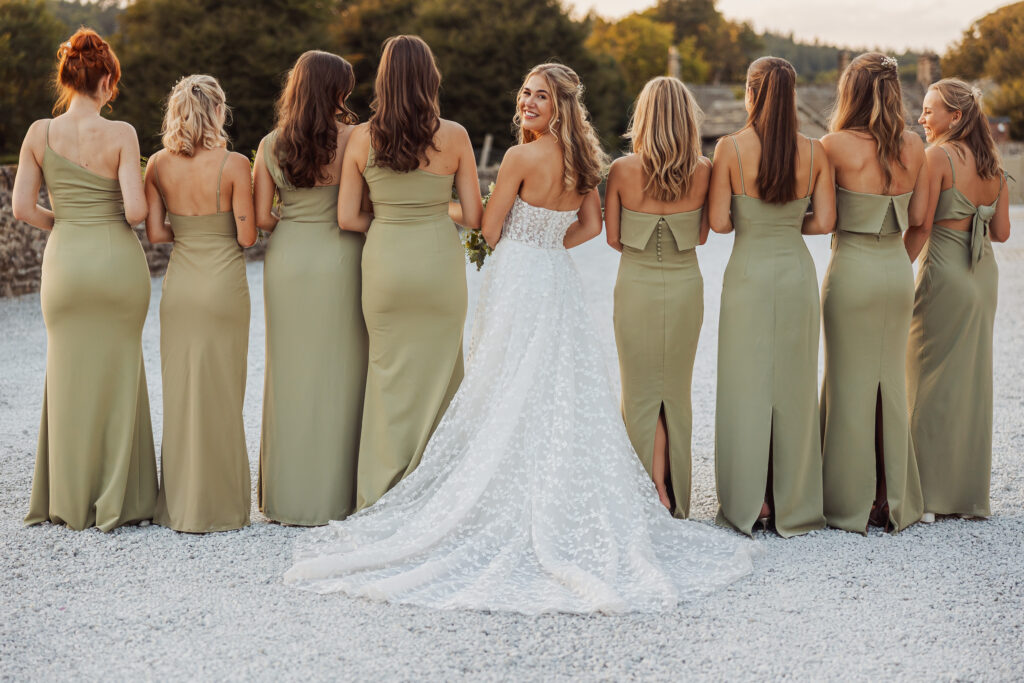 A bride in a white gown stands outdoors at her North Yorkshire wedding with bridesmaids in matching light green dresses, all facing away except the bride, who looks back and smiles at the camera.