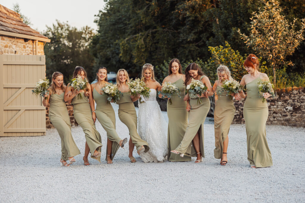 A bride in a white dress and eight bridesmaids in matching sage green dresses stand in a row outdoors at Bolton Abbey, smiling and lifting one leg as they hold bouquets of flowers at this joyful North Yorkshire wedding.