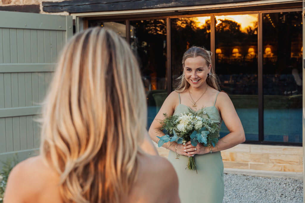 A woman in a light green dress holding a bouquet of flowers smiles at another woman with long blonde hair, standing outdoors near a tithe barn with large windows at sunset during a North Yorkshire wedding.