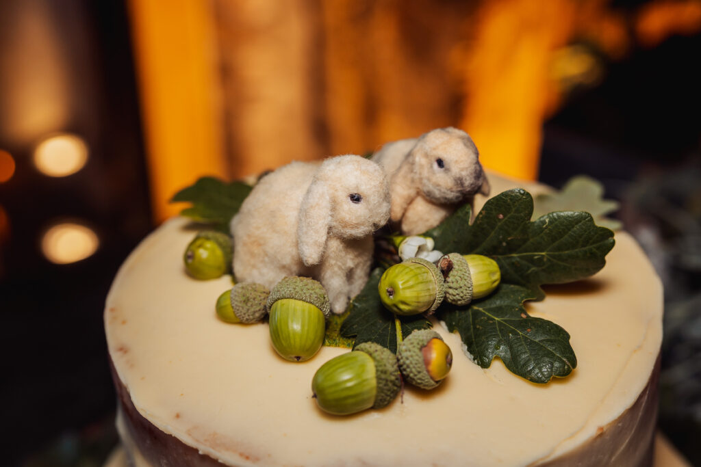 A wedding cake topped with two small plush bunny figures, acorns, and green oak leaves, set against a softly lit background—perfect for a North Yorkshire wedding or a celebration near Bolton Abbey.