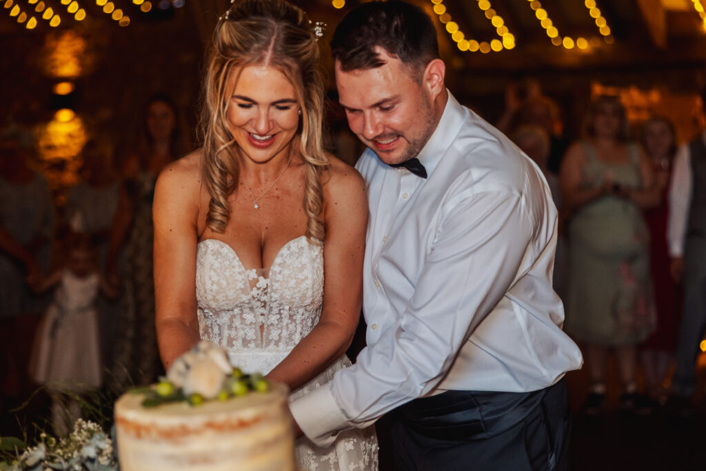A bride and groom, both smiling, cut their wedding cake together at a warmly lit tithe barn wedding reception near Bolton Abbey, with guests and string lights in the background.