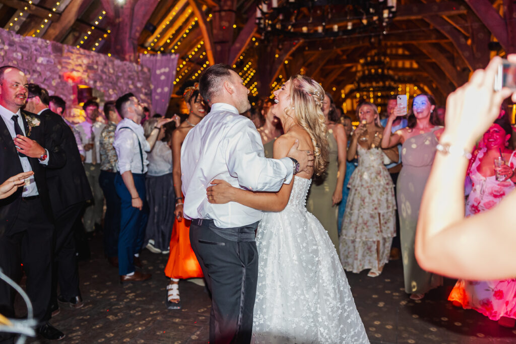 A bride and groom dance together at a lively Bolton Abbey wedding reception, surrounded by guests in formal attire inside a rustic, warmly lit venue decorated with string lights.