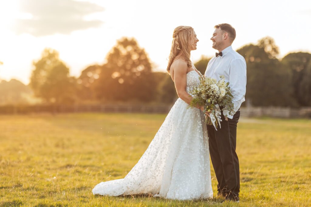 A bride in a white dress and a groom in a black bow tie stand smiling in a sunlit field at their North Yorkshire wedding, holding a bouquet, with trees and the wooden fence of Tithe Barn, Bolton Abbey in the background.