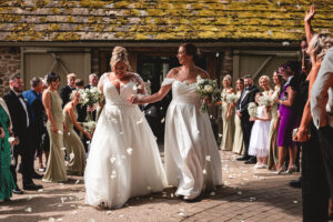 Two brides in white dresses walk hand in hand smiling, surrounded by guests throwing flower petals—the best wedding confetti—outside a rustic building with a mossy roof during a joyful celebration.