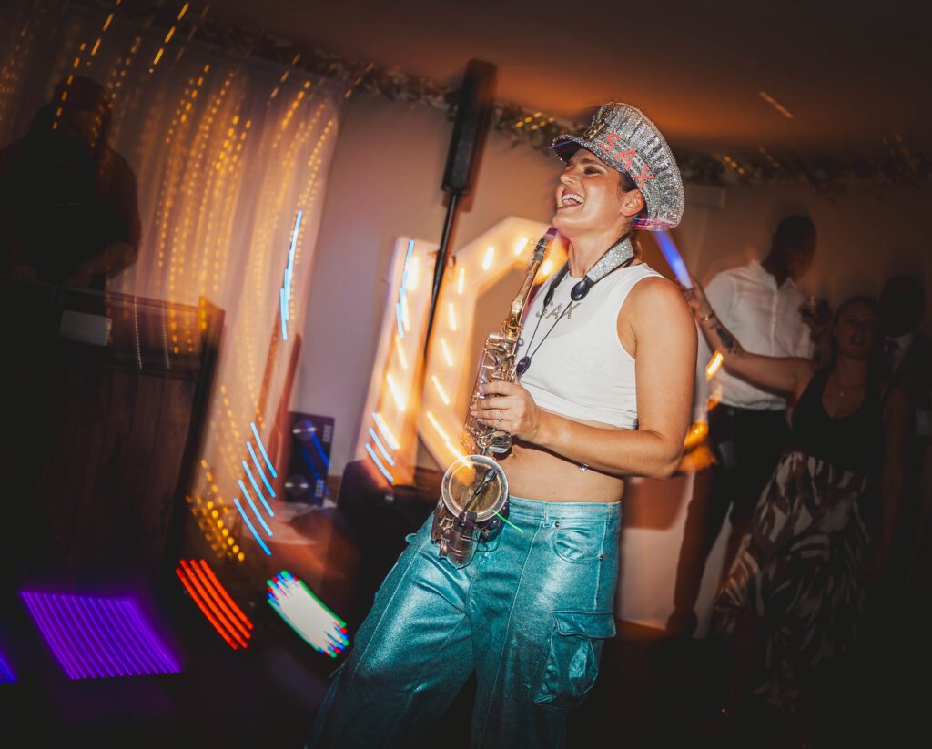 Ellie_Sax_Wedding (12) A woman in a sparkling hat and shiny blue pants smiles while holding a saxophone at a lively indoor party, channeling Ellie Sax vibes as colorful lights dance around blurred partygoers in the background.
