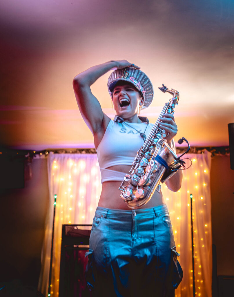 Ellie_Sax_Wedding (4) A woman in a shiny hat and crop top holds a saxophone, posing energetically on stage with colorful lights and a curtain of string lights behind her.