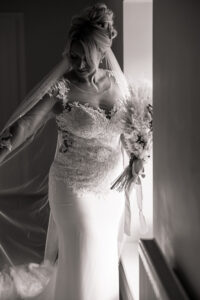 A bride in a lace wedding dress holds a bouquet while standing by a window at Hazel Gap Barn. Light gently illuminates her as she looks down, her veil draping over her arm. The photo, taken at this stunning Cripps and Co venue, is in black and white.