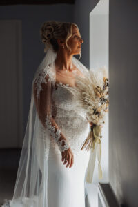 A bride in a lace wedding dress and veil stands by a window at Hazel Gap, holding a bouquet of dried flowers and gazing into the soft natural light, capturing the romantic charm of a Hazel Gap Barn wedding.
