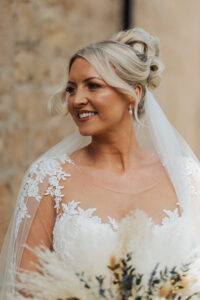 A smiling bride with blonde hair in an elegant updo wears a lace wedding dress and a veil, holding a bouquet of dried flowers, standing in front of a stone wall at her romantic Hazel Gap Barn wedding.
