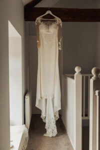 A white lace wedding dress with long sheer sleeves hangs on a hanger in a bright hallway at Hazel Gap Barn, with white railings and a wooden beam above—perfect for a romantic Hazel Gap Barn wedding.