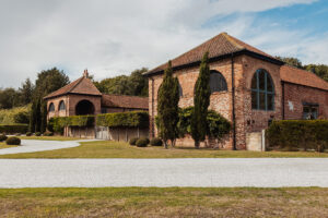 A large brick building with arched windows and a red-tiled roof, Hazel Gap Barn by Cripps and Co is surrounded by trimmed hedges, small round bushes, and tall trees under a partly cloudy sky. A gravel path curves in front of the barn.
