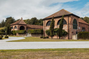 Two rustic brick buildings with arched windows and tiled roofs sit beside a white gravel driveway at Hazel Gap Barn, surrounded by manicured lawns, bushes, and tall trees under a partly cloudy sky—perfect for a Cripps and Co wedding.