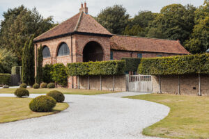 A brick house with a tiled roof and arched windows sits amid neatly trimmed hedges, much like venues by Cripps and Co. A white gravel driveway winds through the well-kept lawn, evoking the scenic charm of a Hazel Gap Barn wedding.