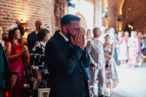 A man in a suit stands indoors, covering his face with his hands in an emotional moment at a Hazel Gap Barn wedding, surrounded by well-dressed guests at this stunning Cripps and Co venue.