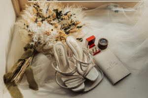 A pair of white strappy heels, a bouquet of dried flowers, a red ring box, and a cream invitation are beautifully arranged on a veiled surface near the window—perfect details for a Hazel Gap wedding inspired by Cripps and Co elegance.