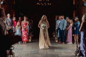 A woman in a beige dress walks down the aisle at Hazel Gap Barn, holding a bouquet, surrounded by guests in formal attire inside the dimly lit venue with lanterns and a brick wall.
