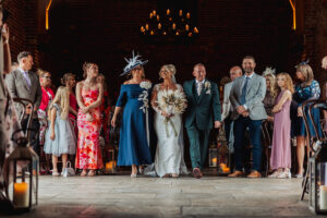 A bride in a white dress walks down the aisle with two people, likely her parents, at a warmly lit Hazel Gap Barn wedding, surrounded by guests in formal and semi-formal attire.