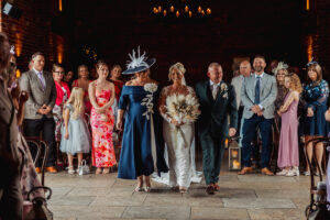 A bride in a white dress walks down the aisle with her parents, surrounded by smiling guests at a warmly lit Hazel Gap Barn wedding by Cripps and Co. Everyone is dressed in formal attire, enhancing the charm of this unforgettable celebration.