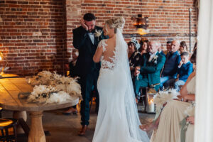 A bride in a white lace gown stands facing a tearful groom during an indoor wedding ceremony at Hazel Gap Barn by Cripps and Co, with guests watching and rustic brick walls in the background.