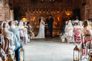 A bride and groom stand at the altar during an indoor Hazel Gap Barn wedding, facing each other and holding hands. Guests are seated in rows, with candles creating a warm, romantic atmosphere in the rustic brick venue.